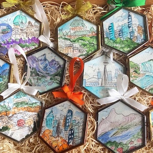May include: A collection of hexagonal ornaments with watercolor cityscapes, each framed in gold and adorned with ribbons. The ornaments depict various landmarks and skylines, showcasing a range of colors and artistic styles. The collection is nestled in a bed of straw.