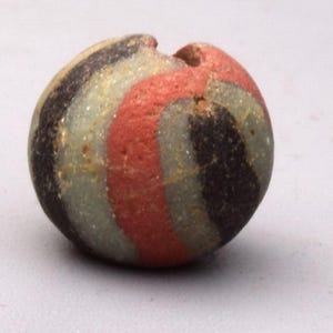 May include: A round, striped bead with a hole. The bead features bands of black, red, and grey. The bead is made of a stone-like material and has a slightly rough texture. The bead is a small, decorative item.