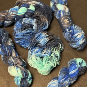 May include: Four skeins of hand-dyed yarn in shades of blue, teal, and gray. The yarn is arranged in a loose pile on a brown surface. The label reads "Bristlecone Company Hand Dyed Yarn".