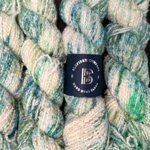 Puede incluir: Madejas de hilo teñido a mano en tonos de azul, verde y blanco. El hilo está retorcido y apilado, con una etiqueta que dice "Blufiber Company Hand Dyed Yarns".
