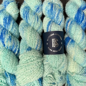 May include: Four skeins of hand-dyed yarn in a light blue and green colorway with a white swirl pattern. The yarn is labeled "Bluefiber Company Hand Dyed Yarns".