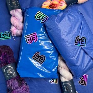 May include: A top-down shot of a blue surface with several skeins of hand-dyed yarn and two wrapped packages. The packages are sealed with holographic stickers featuring a stylized "B" logo. The yarn colors range from purple to pink and yellow.