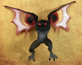 May include: A black bat ornament with red and gold wings. The bat has large red eyes and is hanging from a string.