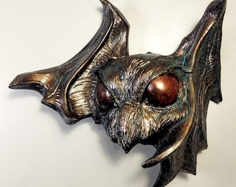 Puede incluir: Una escultura de murciélago de color bronce con alas texturizadas y grandes ojos rojos. La escultura tiene un acabado desgastado, lo que le da un aspecto antiguo.