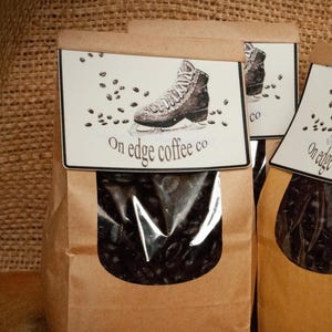Op de afbeelding: Vier bruine papieren zakken met koffiebonen en een doorzichtig venster. Elke zak heeft een label met een tekening van een laars en de tekst "On edge coffee co". De zakken staan op een houten oppervlak.