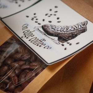 Op de afbeelding: Een bruine papieren koffiezak met een doorzichtig venster met koffiebonen. Het etiket bevat de tekst "On edge coffee co" en een illustratie van een vintage schaats en koffiebonen.