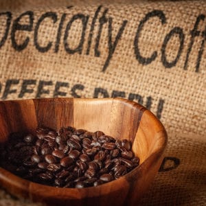 Może przedstawiać: Drewniana miska wypełniona prażonymi ziarnami kawy. Miska stoi przed workiem jutowym z nadrukiem "specialty Coffee". Ziarna kawy są ciemnobrązowe.