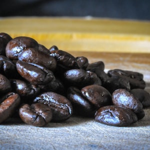 May include: A close-up shot of a pile of roasted coffee beans. The beans are a rich, dark brown color with a glossy sheen, suggesting freshness. The image is well-lit, highlighting the texture and shape of the beans, perfect for coffee lovers.