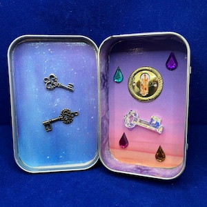 Può includere: Una scatola aperta color argento con uno sfondo sfumato blu e viola. Il lato sinistro ha due piccole chiavi di metallo. Il lato destro presenta un ciondolo a forma di buco della serratura, una chiave di cristallo e diversi abbellimenti a forma di gioiello.
