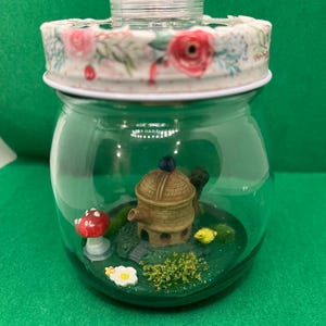 Puede incluir: Un terrario de vidrio transparente con una tapa estampada floral y una parte superior de plástico transparente. En el interior, una escena en miniatura presenta una seta, un edificio y otros pequeños elementos decorativos. El terrario se encuentra sobre una superficie verde.