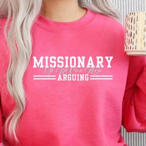 Puede incluir: Una sudadera rosa brillante con el texto blanco "MISSIONARY So We Can Keep ARGUING". La sudadera es de cuello redondo. Una taza blanca con un dise&ntilde;o dorado se sostiene en la mano derecha.