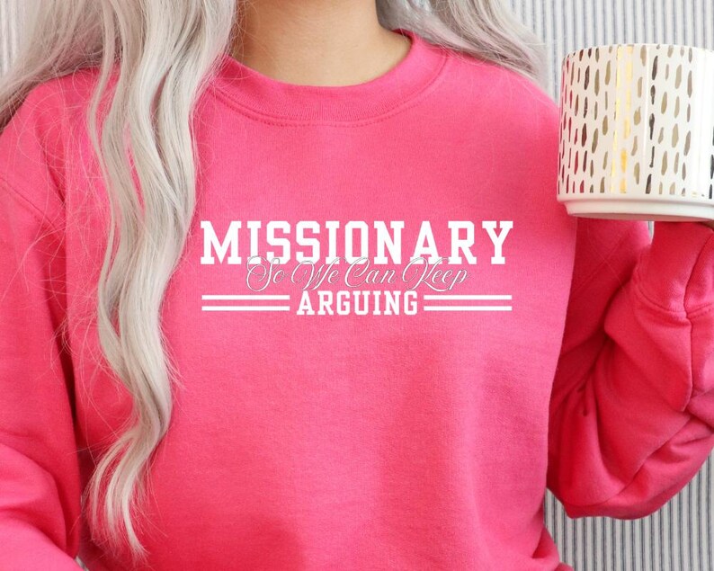 Puede incluir: Una sudadera rosa brillante con el texto blanco "MISSIONARY So We Can Keep ARGUING". La sudadera es de cuello redondo. Una taza blanca con un dise&ntilde;o dorado se sostiene en la mano derecha.