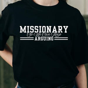 Puede incluir: Camiseta negra de cuello redondo con el texto blanco "MISSIONARY So We Can Keep Arguing". El texto est&aacute; en negrita, con una fuente decorativa para "So We Can Keep". Dos l&iacute;neas horizontales est&aacute;n debajo de la palabra "ARGUING". Una prenda casual.