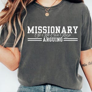 Puede incluir: Camiseta gris oscuro con el texto blanco "MISSIONARY So We Can Keep ARGUING". La camiseta tiene cuello redondo y mangas cortas. La tela parece suave y c&oacute;moda. Una prenda informal para todos los d&iacute;as.