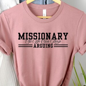 Puede incluir: Camiseta rosa polvoriento con el texto negro &laquo;MISSIONARY So We Can Keep ARGUING&raquo;. El texto est&aacute; centrado y enmarcado por dos l&iacute;neas horizontales. La camiseta est&aacute; colgada de una percha de madera.