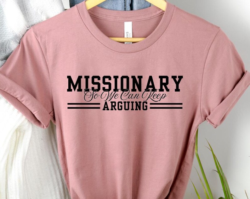 Puede incluir: Camiseta rosa polvoriento con el texto negro &laquo;MISSIONARY So We Can Keep ARGUING&raquo;. El texto est&aacute; centrado y enmarcado por dos l&iacute;neas horizontales. La camiseta est&aacute; colgada de una percha de madera.