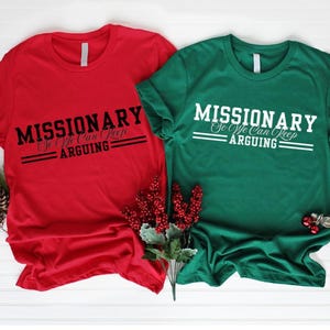 Puede incluir: Dos camisetas de manga corta, una roja y otra verde, con el texto "MISSIONARY So We Can Keep Arguing" impreso en blanco y negro. Las camisetas se muestran sobre una superficie blanca con adornos navide&ntilde;os.