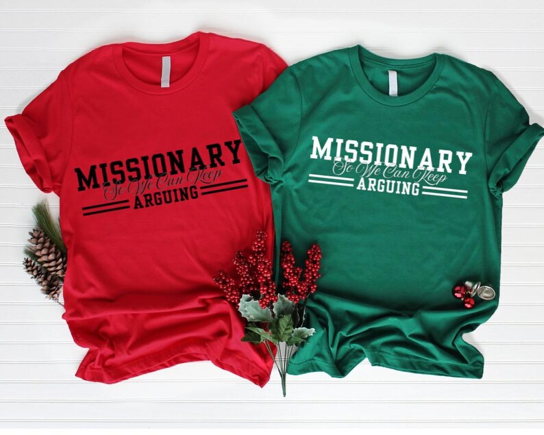 Puede incluir: Dos camisetas de manga corta, una roja y otra verde, con el texto "MISSIONARY So We Can Keep Arguing" impreso en blanco y negro. Las camisetas se muestran sobre una superficie blanca con adornos navide&ntilde;os.