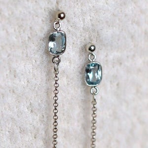 May include: A pair of silver drop earrings featuring a small, polished silver ball stud at the top, connected to a rectangular, light blue faceted gemstone in a silver bezel setting, with a delicate silver chain hanging from the bottom of each stone.