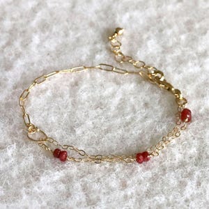 May include: A delicate gold-toned bracelet featuring a double chain design. Small, vibrant red beads are evenly spaced along the lower chain. The bracelet has a lobster clasp closure and is displayed on a white, textured surface.