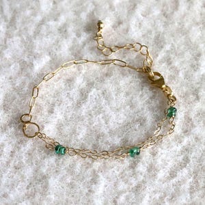 Peut inclure: Un bracelet doré délicat avec de petits maillons ovales et une série de petites pierres précieuses vertes. Le bracelet a une chaîne réglable et une fermeture à fermoir. Le bijou est présenté sur une surface blanche texturée.