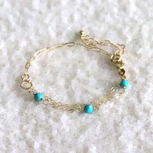 Könnte beinhalten: Ein zartes Armband aus goldfarbener Kette mit zwei kleinen, runden, leuchtend türkisfarbenen Perlen, die entlang der Glieder verteilt sind. Das Armband verfügt über eine feine Goldkette mit Karabinerverschluss und eine verstellbare Verlängerungskette.