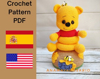 May include: Crochet pattern PDF for a Winnie the Pooh amigurumi doll. The doll is yellow with a red shirt and is sitting on a brown crocheted base with bees. The pattern is available in both English and Spanish.