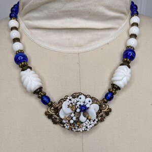 Puede incluir: Un collar de cuentas de vidrio blanco y azul con un gran colgante floral blanco y dorado.