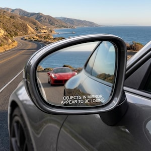 May include: A grey car side mirror with a reflective surface showing a red sports car on a coastal road. The mirror glass features the white printed text: OBJECTS IN MIRROR APPEAR TO BE LOSING. The mirror housing is smooth and curved in design.