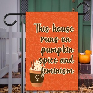 Peut inclure: Drapeau de jardin orange avec un motif de citrouilles et de feuilles. Le drapeau porte le texte "This house runs on pumpkin spice and feminism" avec un dessin de tasse à café et un symbole féminin.