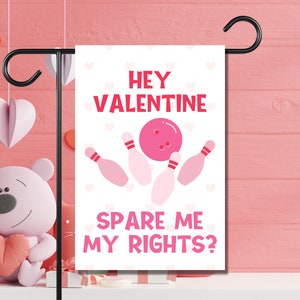 以下が含まれることがあります： ボウリングボールとピンが描かれたピンクと白のバレンタインデーのガーデンフラッグ。フラッグには「Hey Valentine Spare Me My Rights?」と書かれています。