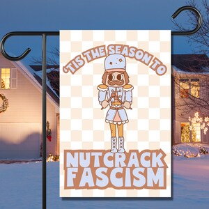 Może przedstawiać: Festiwalna choinka ogrodowa z kreskówkową ilustracją postaci łamacza orzechów trzymającego bęben. Flaga ma szachownicowe tło i napis "Tis the season to Nutcrack Fascism".
