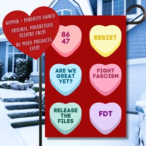 Può includere: Una bandiera da giardino rossa con caramelle a forma di cuore. Le caramelle hanno frasi come "RESIST" e "FIGHT FASCISM". Un cuore rosso con il testo "WOMAN + MINORITY OWNED ORIGINAL PROGRESSIVE DESIGNS ONLY! NO MAGA PRODUCTS EVER!" è in alto.