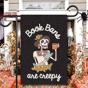 May include: A black garden flag featuring a skeleton holding a book and a mug. The text on the flag reads "Book Bans are creepy".
