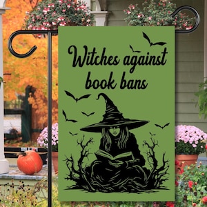 May include: A green garden flag with the text "Witches against book bans" in black script. The flag features a black silhouette of a witch reading a book, surrounded by bats and stylized trees.