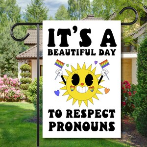Peut inclure: Un drapeau de jardin blanc avec un soleil jaune souriant et des drapeaux arc-en-ciel. Le texte sur le drapeau dit "C'est une belle journée pour respecter les pronoms".