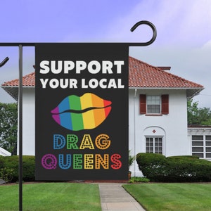 May include: A black garden flag with the text "Support Your Local Drag Queens" in white. A rainbow-coloured lips design is above the text.