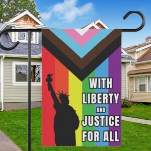 May include: A colourful garden flag with a rainbow design and the text "With Liberty and Justice for All". The flag features a silhouette of the Statue of Liberty holding a torch.