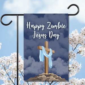 Könnte beinhalten: Eine Gartenflagge mit dem Text "Happy Zombie Jesus Day" in weißer Schrift. Die Flagge zeigt ein Holzkreuz mit einem hellblauen Tuch vor einem bewölkten blauen Hintergrund. Die Flagge wird auf einem schwarzen Metallständer präsentiert, mit weißen Blumen und blauem Himmel im Hintergrund.