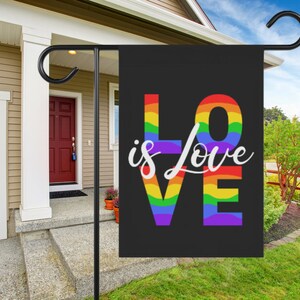 May include: A black garden flag with the word "LOVE" in rainbow colors and the words "is love" in white text.