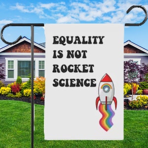May include: A white garden sign with the text "Equality is not rocket science" in black letters. A rainbow rocket ship is pictured below the text.