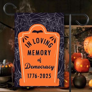 May include: A Halloween garden flag with a tombstone design. The orange tombstone reads "In Loving Memory of Democracy 1776-2025" against a dark blue background with spiderwebs. Pumpkins and a black metal stand are also visible.