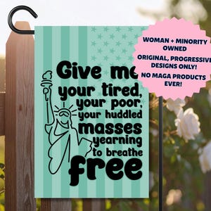 May include: A garden flag with a black metal hanger. The flag has a light teal background with vertical stripes and stars. It features a black graphic of the Statue of Liberty and the text "Give me your tired, your poor, your huddled masses yearning to breathe free."