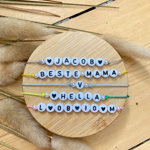 Könnte beinhalten: Vier bunte Freundschaftsarmbänder mit weißen Perlen und schwarzen Buchstaben, die "JACOB", "BESTE MAMA", "HELLA" und "LOVE" buchstabieren.