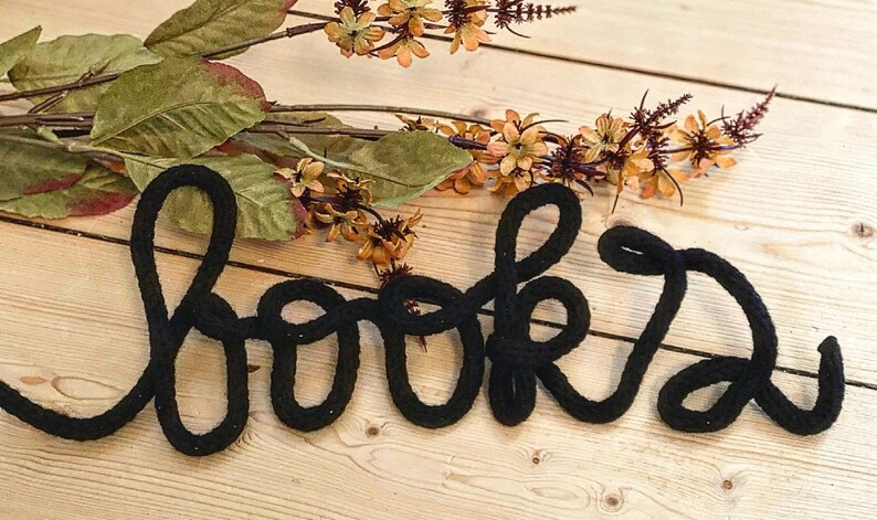 May include: Black yarn word art spelling out "books" on a light wood background.
