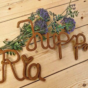 May include: The names "Ria" and "Jupp" spelled out in brown yarn on a wooden surface with purple flowers.