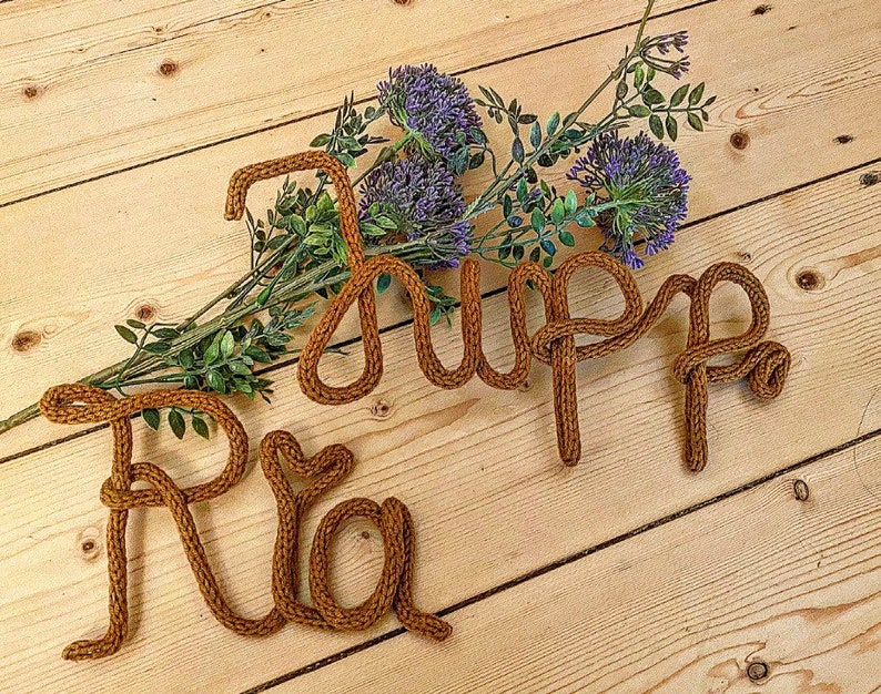 May include: The names "Ria" and "Jupp" spelled out in brown yarn on a wooden surface with purple flowers.