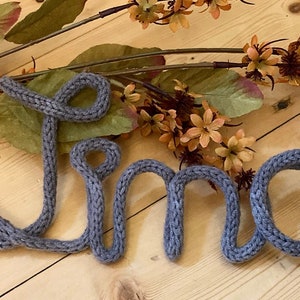 May include: The name "Lino" is spelled out in gray yarn, with each letter formed in a loop. The yarn is knitted and has a textured appearance. The name is lying on a light wood surface.