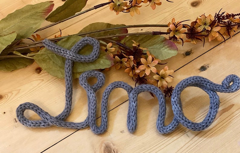 May include: The name "Lino" is spelled out in gray yarn, with each letter formed in a loop. The yarn is knitted and has a textured appearance. The name is lying on a light wood surface.