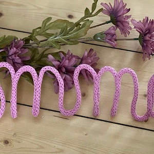 May include: Pink knitted yarn spelling out the name "Emma" on a light wood background with a sprig of purple flowers.